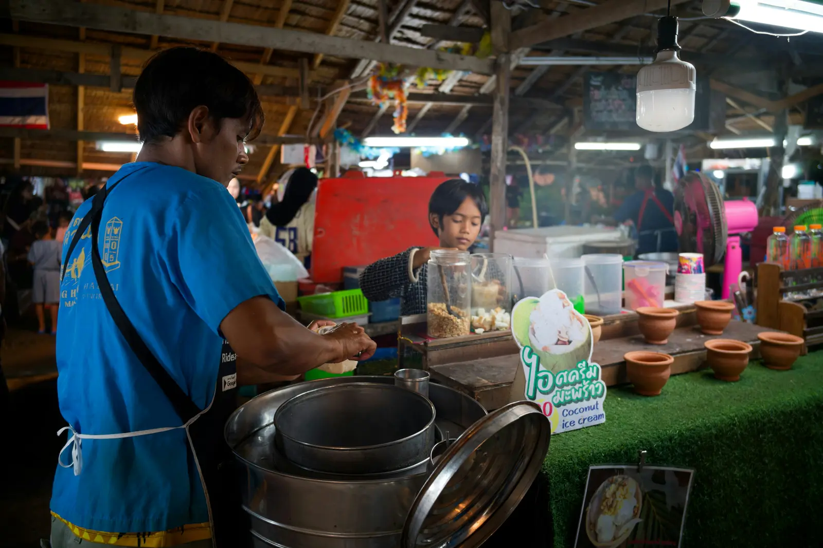 แม่ค้าขายไอศกรีมกะทิในตลาดนัดไทย มีเด็กช่วยตักท็อปปิ้งอยู่หลังร้าน
